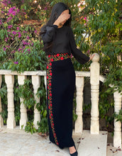 Woman wearing a black outfit with floral embroidery standing outdoors near a white railing.